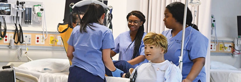 Two nursing students attend to a patient using a lift device, while another student wheels a mannequin patient in a wheelchair.