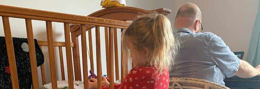 A toddler plays with a toy next to her crib in the foreground while her dad is working on his laptop in the background
