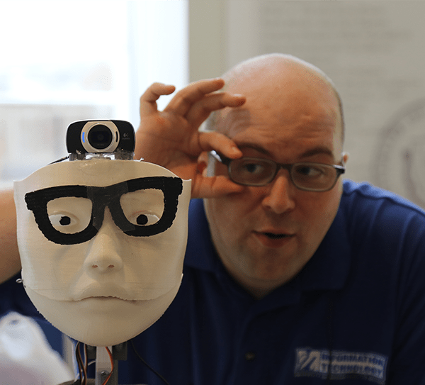 John Mazzarella appears beside a 3D creation, a human like face with glasses and a video camera, in the MakerSpace