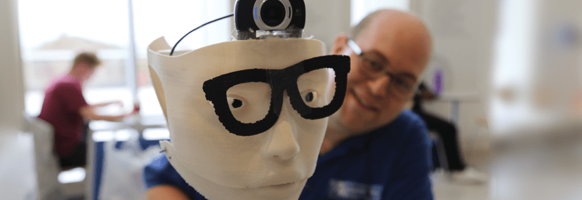 John Mazzarella appears smiling behind a 3D creation, a human like face with glasses and a video camera, in the MakerSpace